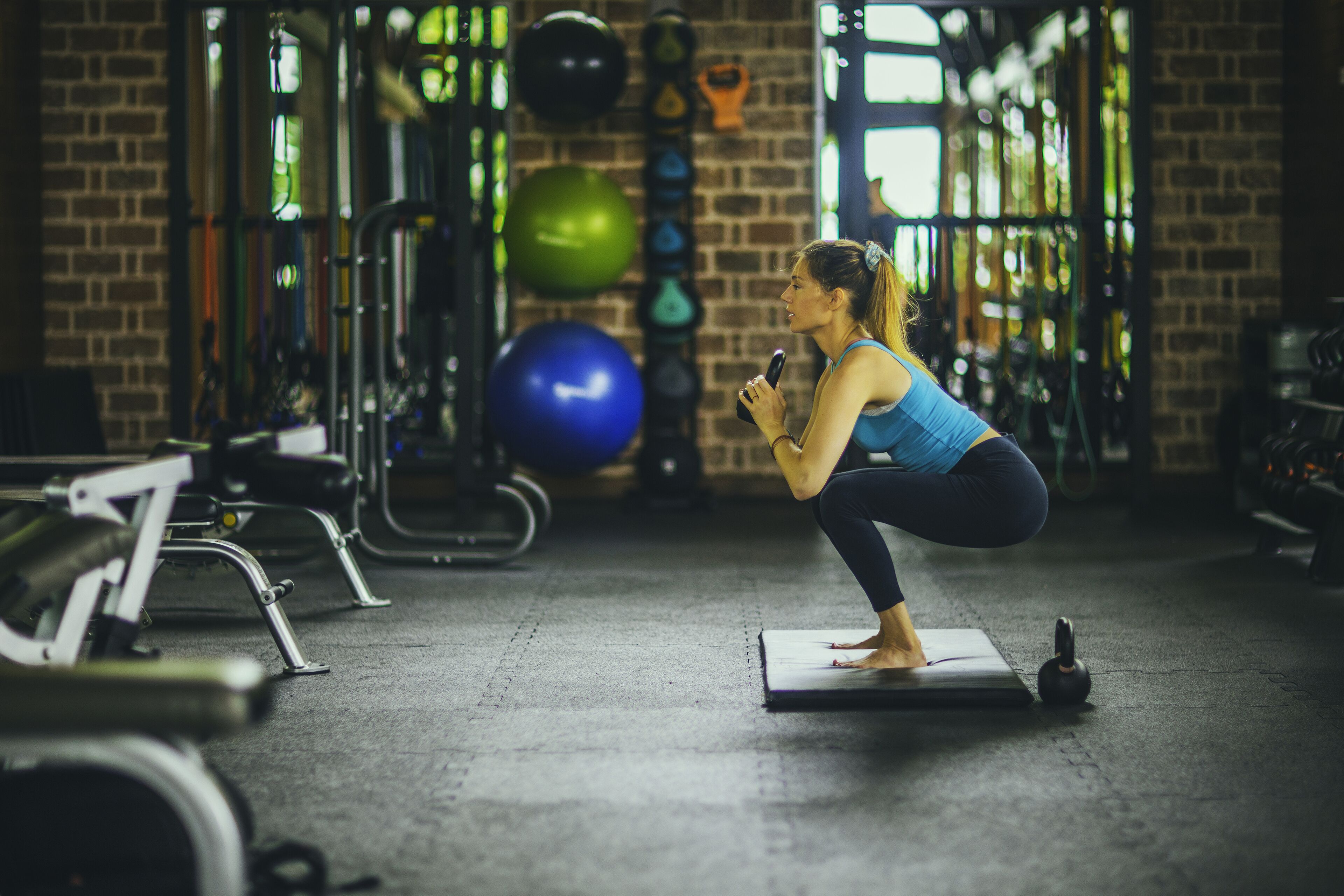 A woman performing squats on a mat in a gym with exercise equipment and a brick wall background.