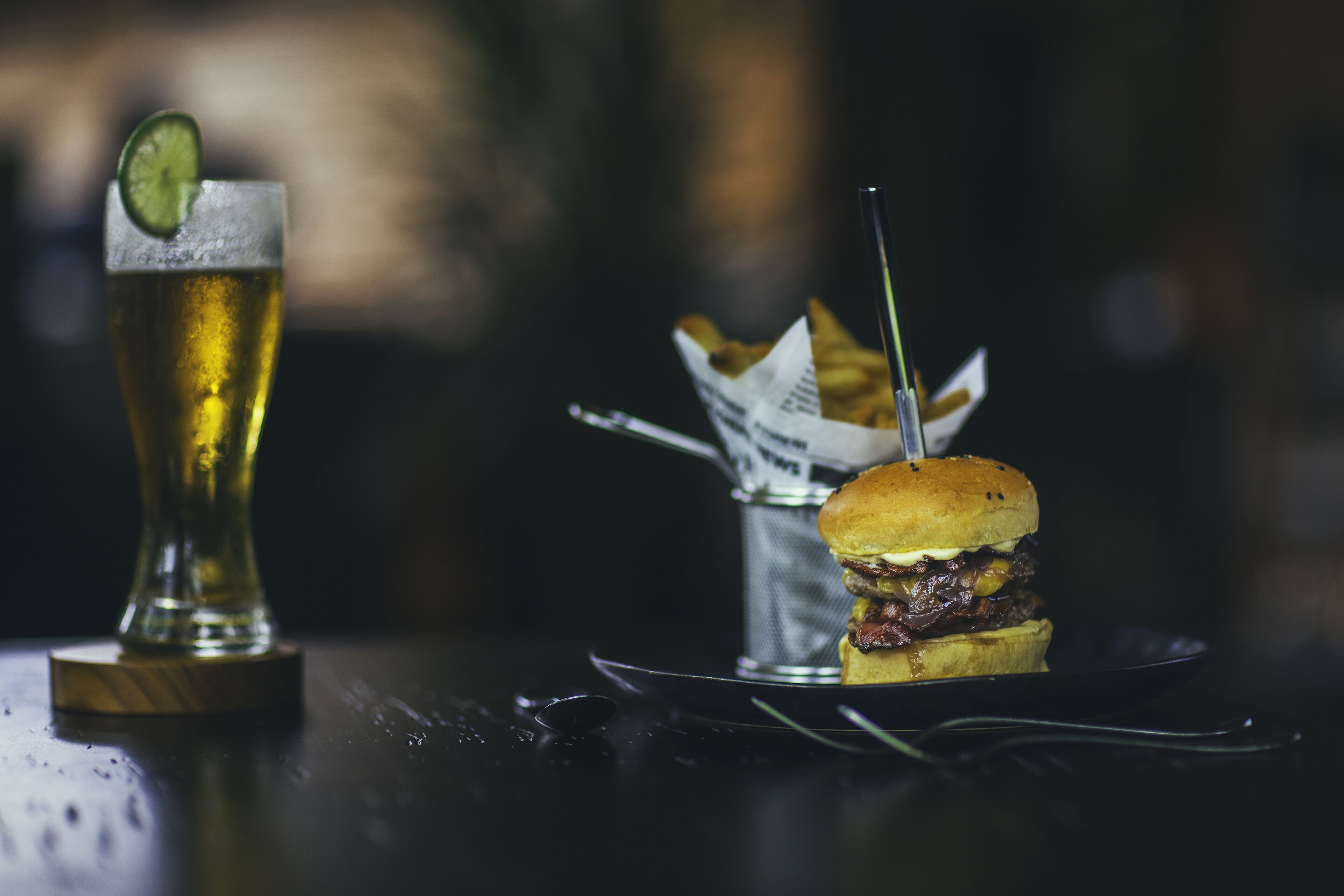A glass of beer with a lime wedge, a burger, and a small container of fries on a table.