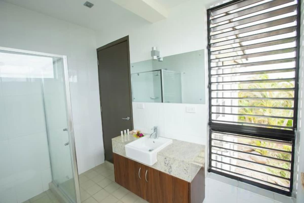 A bathroom with a glass shower enclosure, a sink with a wooden vanity, and a window with horizontal blinds.