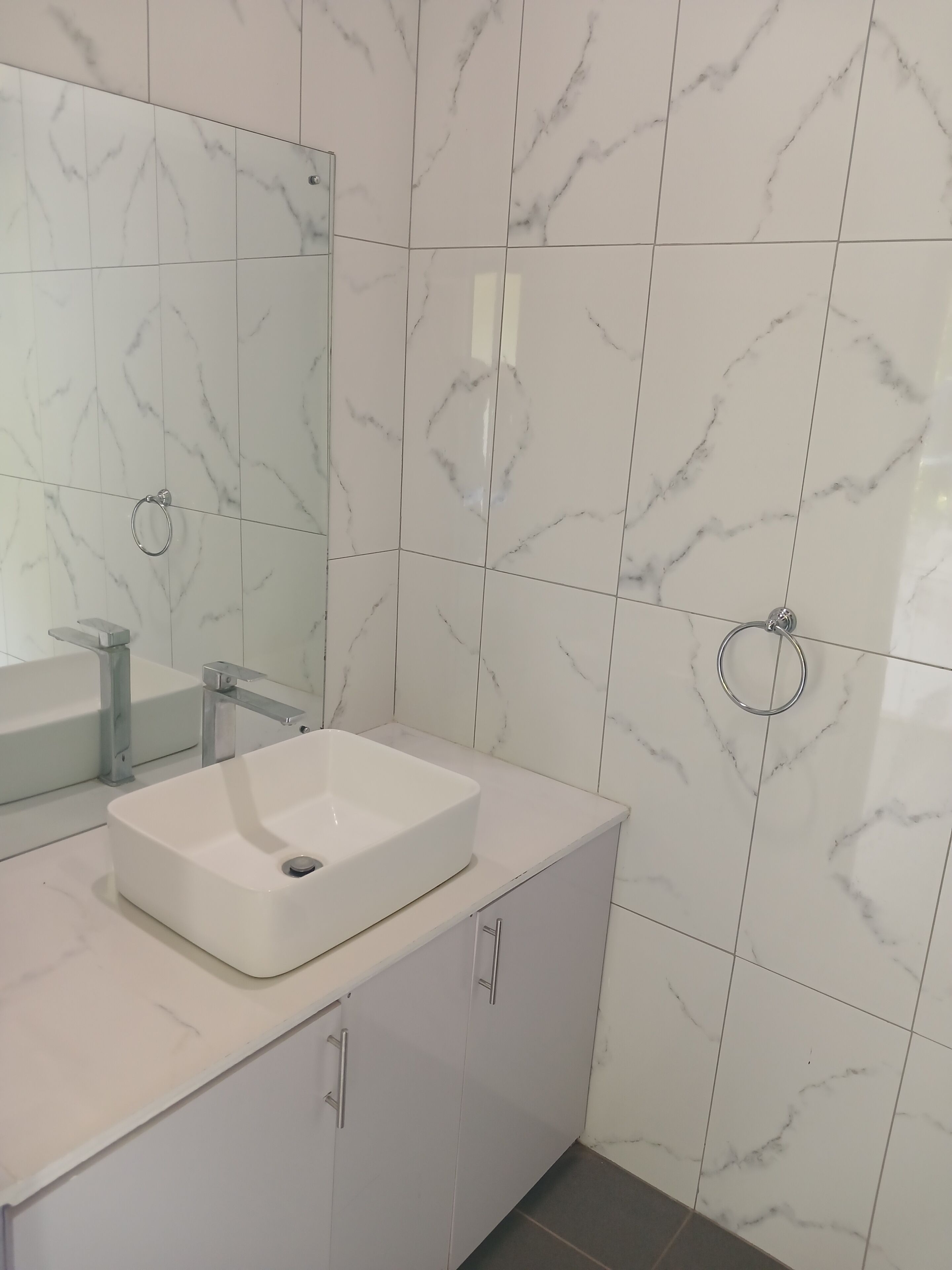 Ein Badezimmer mit weißen Marmorfliesen, einem weißen Waschbecken, einem Spiegel und einem Handtuchhalter.