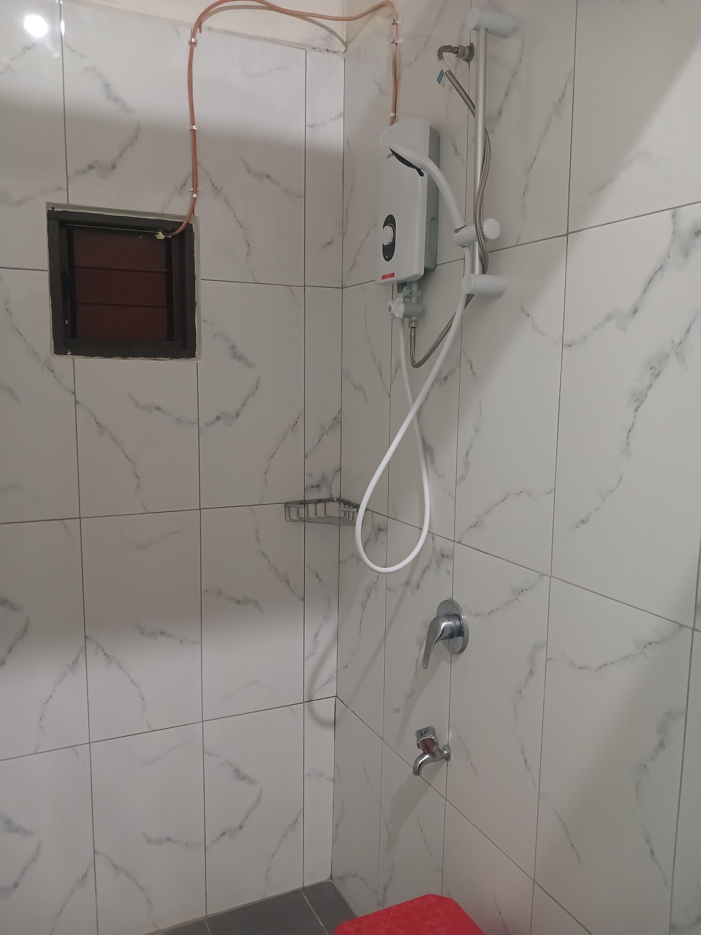 A bathroom with white marble tiles, a shower with a handheld showerhead, and a small window with a brown frame.