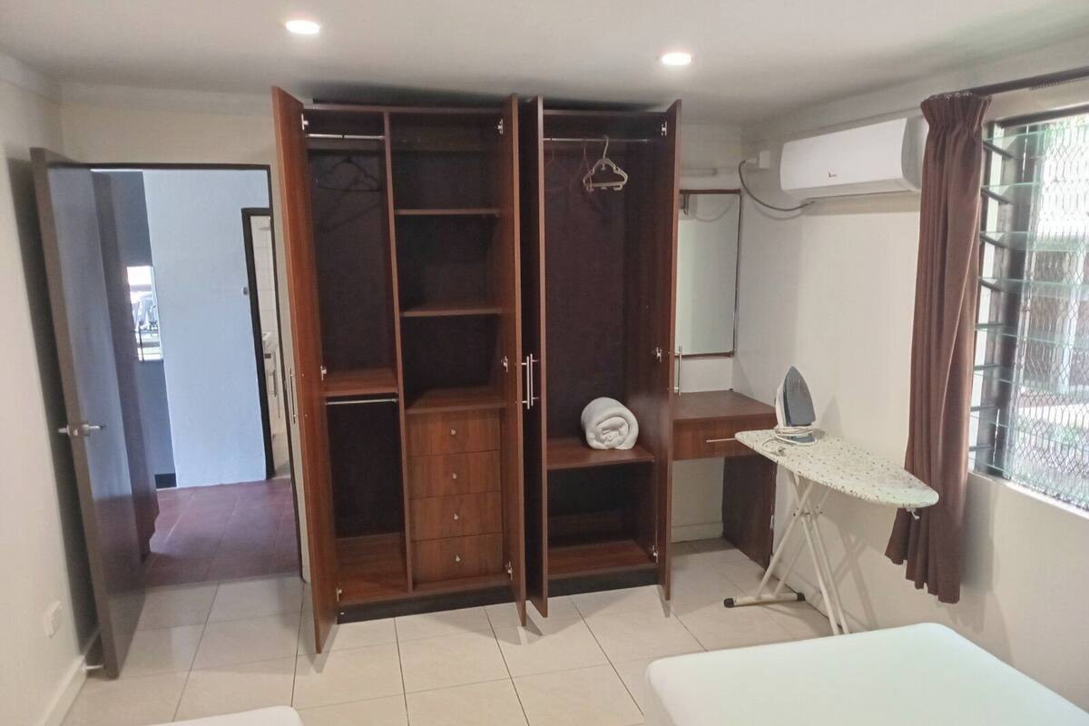 A bedroom with a wardrobe, a bedside table with an iron, a window with curtains, and a door leading to another room.