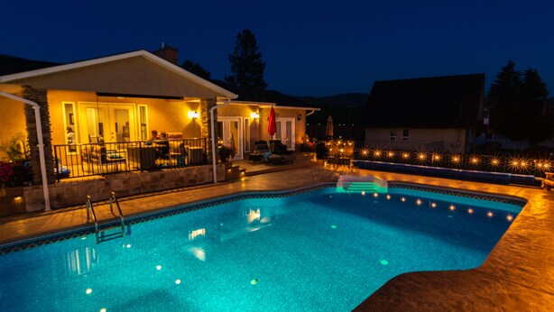 Una piscina con un área de patio techada, una escalera y una terraza bien iluminada.