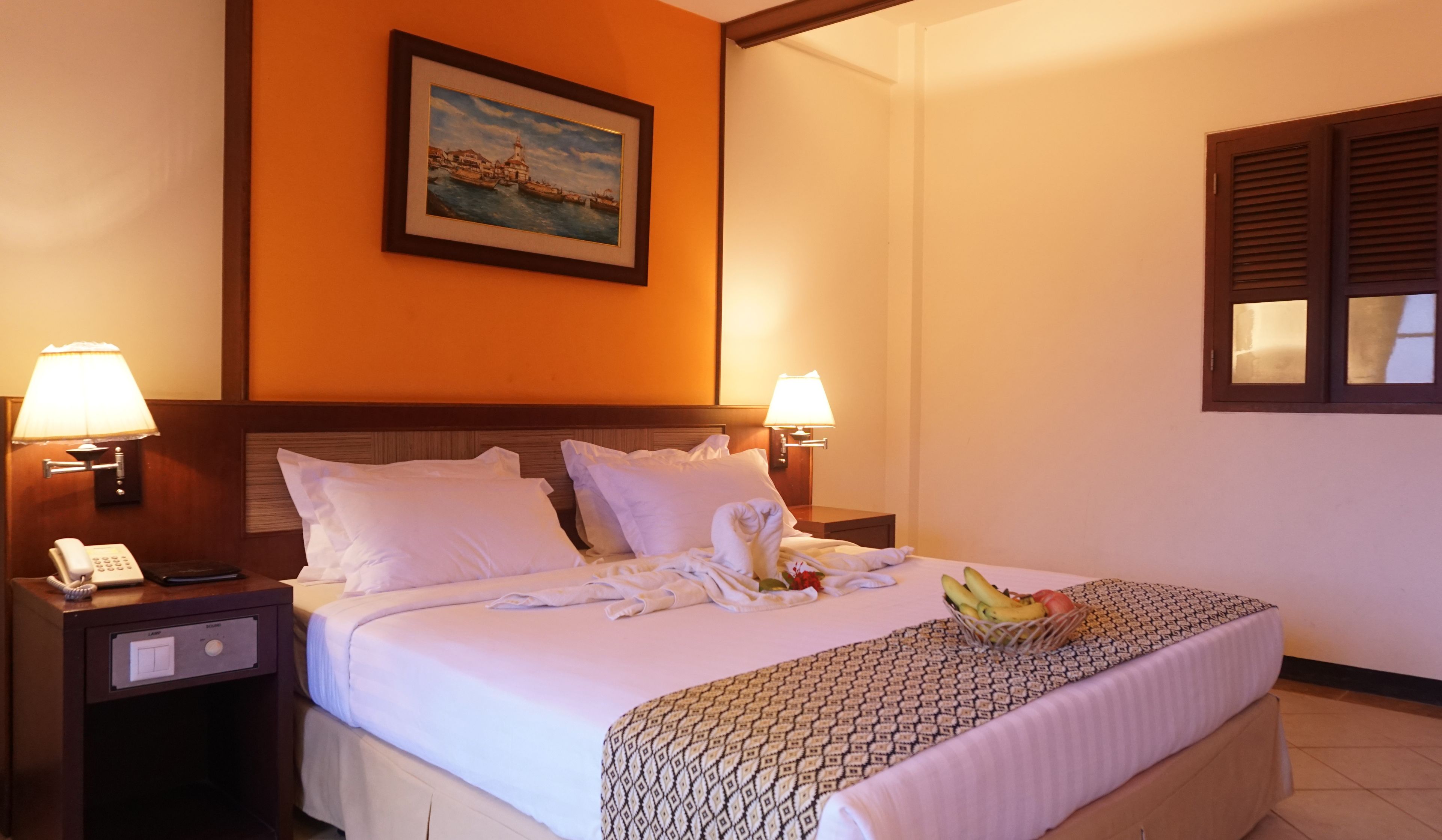 A hotel room with a bed, two bedside lamps, a nightstand with a phone, a painting on the wall, and a window with shutters.