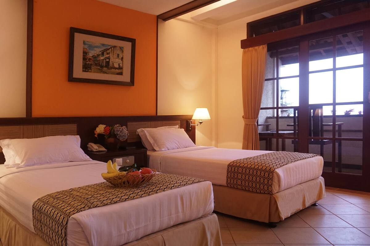 Una camera d'albergo con due letti, un comodino con una lampada, un quadro appeso al muro e una finestra con tende.