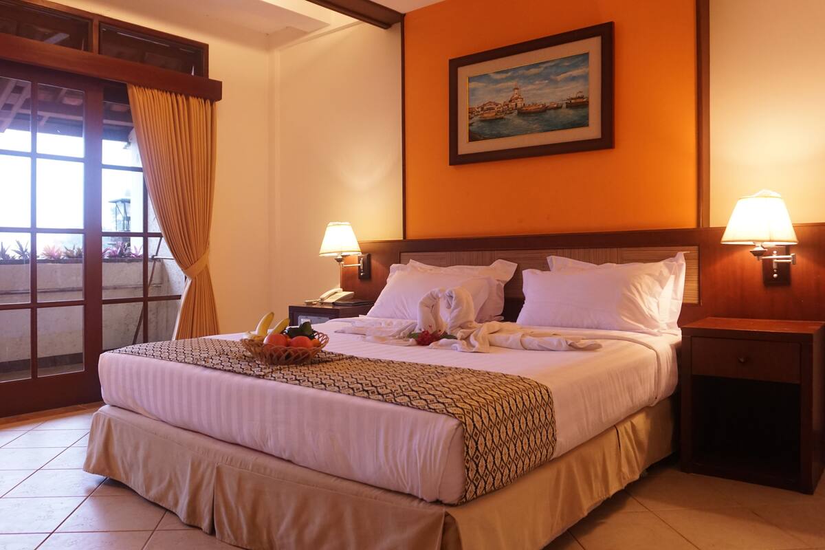 Una camera d'albergo con un letto grande, due comodini con lampade, un quadro al muro e una vista attraverso una finestra con tende.