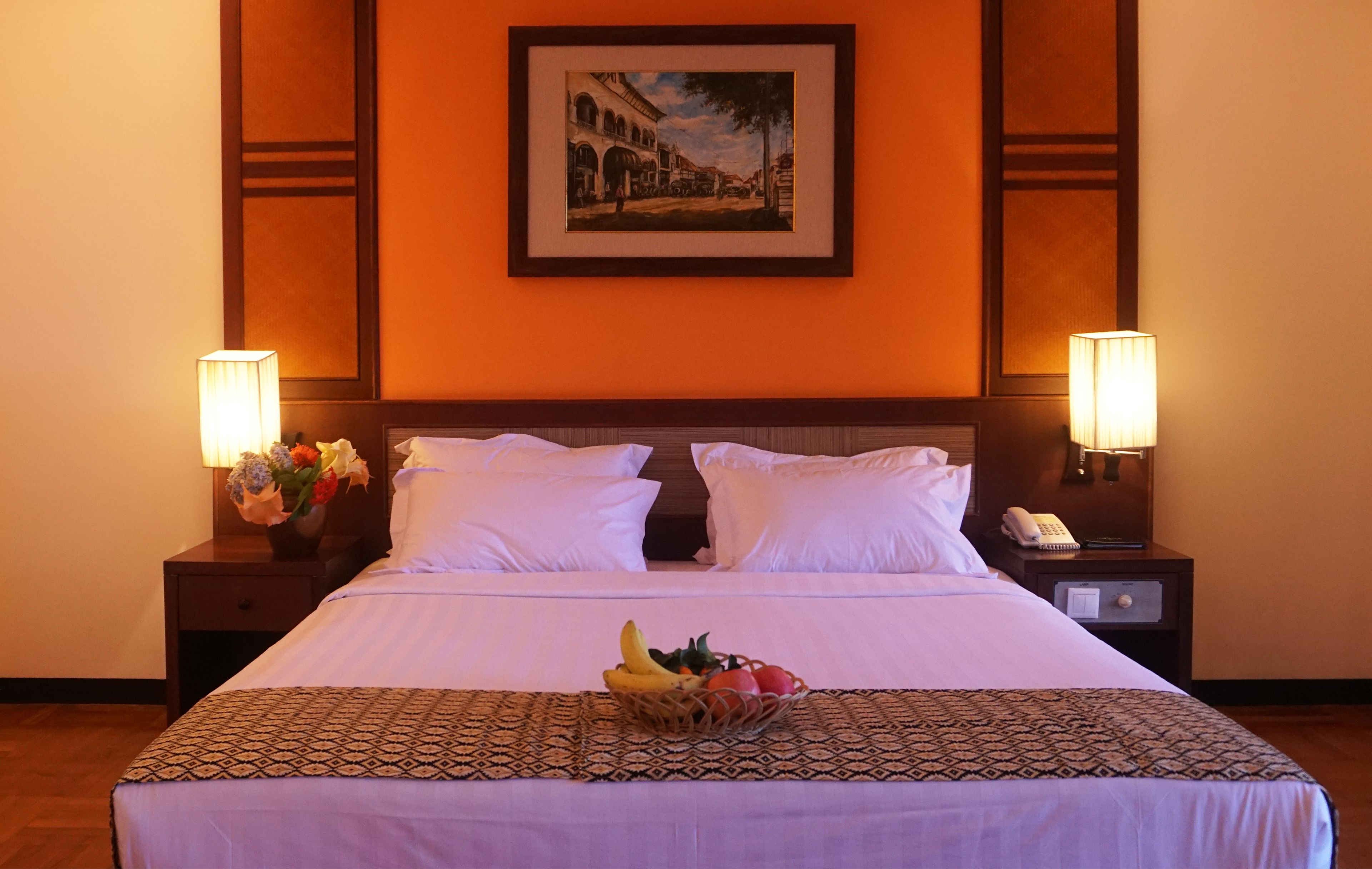 Une chambre d’hôtel avec un grand lit, deux lampes de chevet, un tableau encadré au mur et un panier de fruits posé sur le lit.