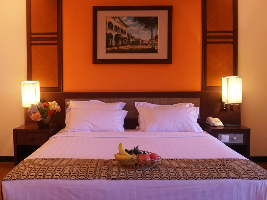Une chambre d’hôtel avec un grand lit, deux lampes de chevet, un tableau encadré au mur et un panier de fruits posé sur le lit.