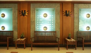 A lobby with three wooden benches, each with a small table and a potted plant. The walls have decorative wall sconces and a large window with a grid pattern. - Pines Garden Resort (Prigen)