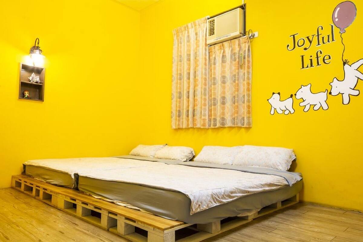 Quarto com cama sobre plataforma de madeira, ar condicionado fixado na parede, decoração com a frase 'Joyful Life' e um balão, e uma pequena estante com uma figurina.