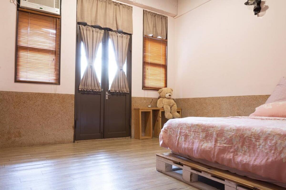 Quarto com cama, mesa de cabeceira de madeira, um urso de pelúcia e porta dupla com cortinas.