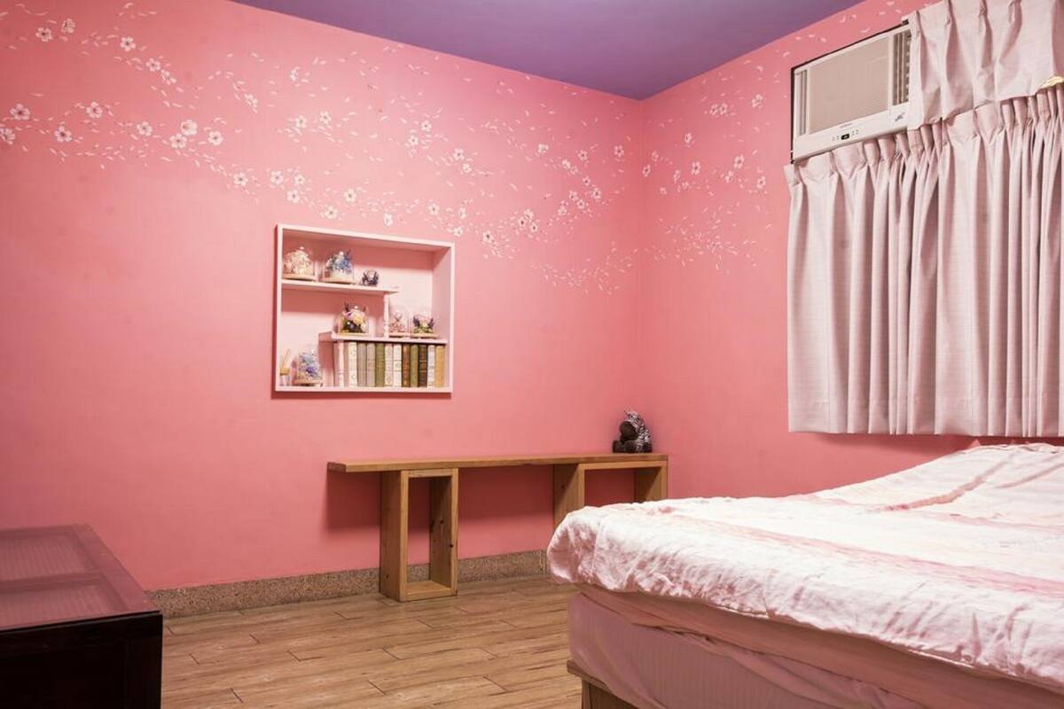 Um quarto rosa com uma cama, uma estante de madeira com livros e objetos decorativos, e uma janela com cortinas.