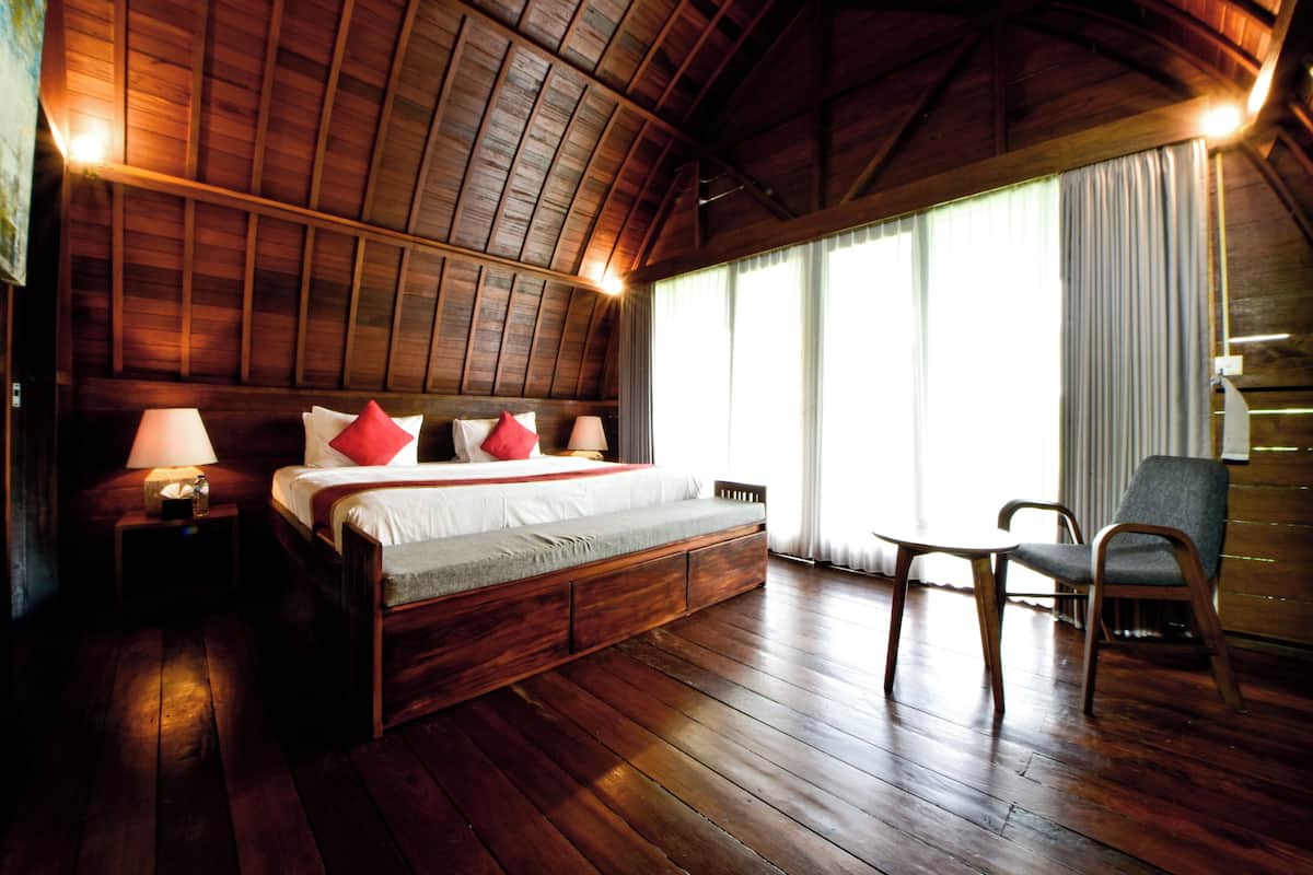 Quarto com piso de madeira, uma cama, abajures, uma cadeira e uma mesinha.