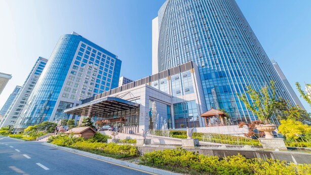 A modern building with a curved glass facade, a covered entrance, and a fountain in the foreground. - Hundred Hotel (Qingdao)