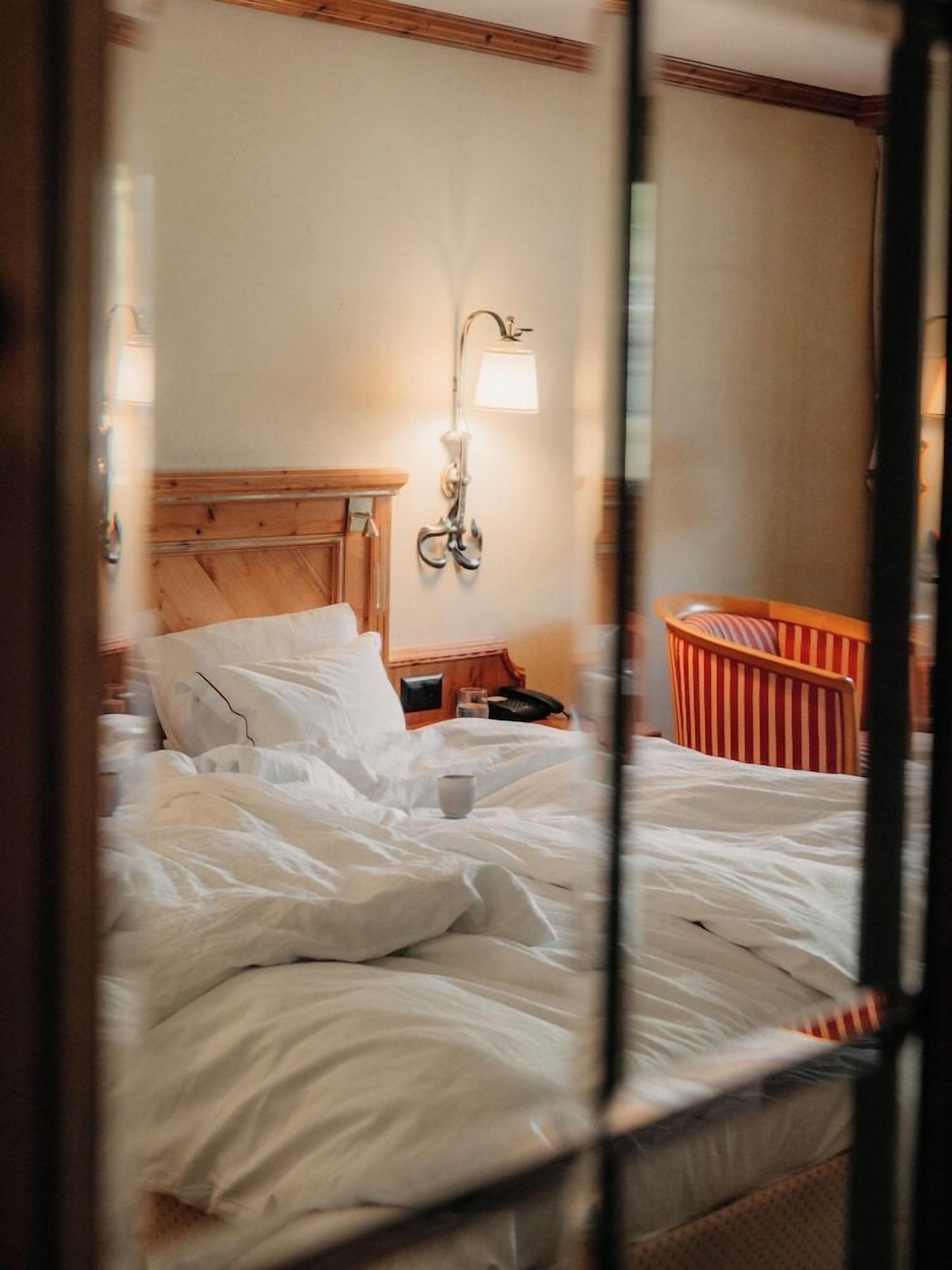 A hotel room with a wooden headboard, a bedside lamp, a striped chair, and a bedside table with a phone.