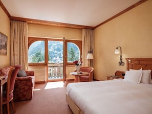A hotel room with a bed, a desk, a chair, a small table with a vase, and a view of mountains through large windows.