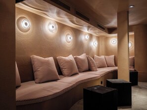 A modern, well-lit seating area with a series of circular light fixtures on the wall, a cushioned bench with striped pillows, and a dark square table.