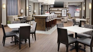 A modern hotel lobby with a central bar area, multiple seating options including tables and chairs, and a large television screen.