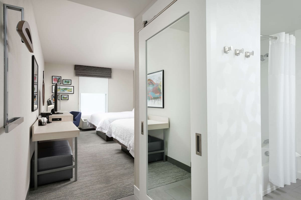 A modern hotel room with a desk, two beds, a shower area, and a view of the city.