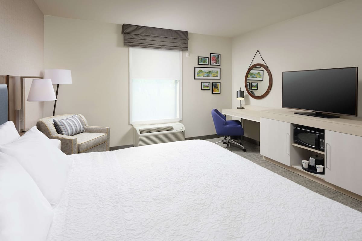 A hotel room with a large bed, a desk with a computer, a chair, a television, and a window with blinds.
