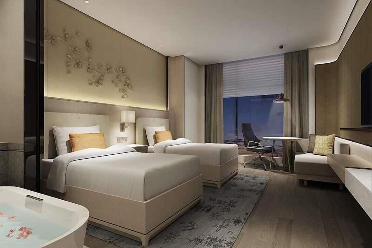 A modern hotel room with a large bed, a sitting area with a sofa and armchairs, a small table, and a large window with a view.