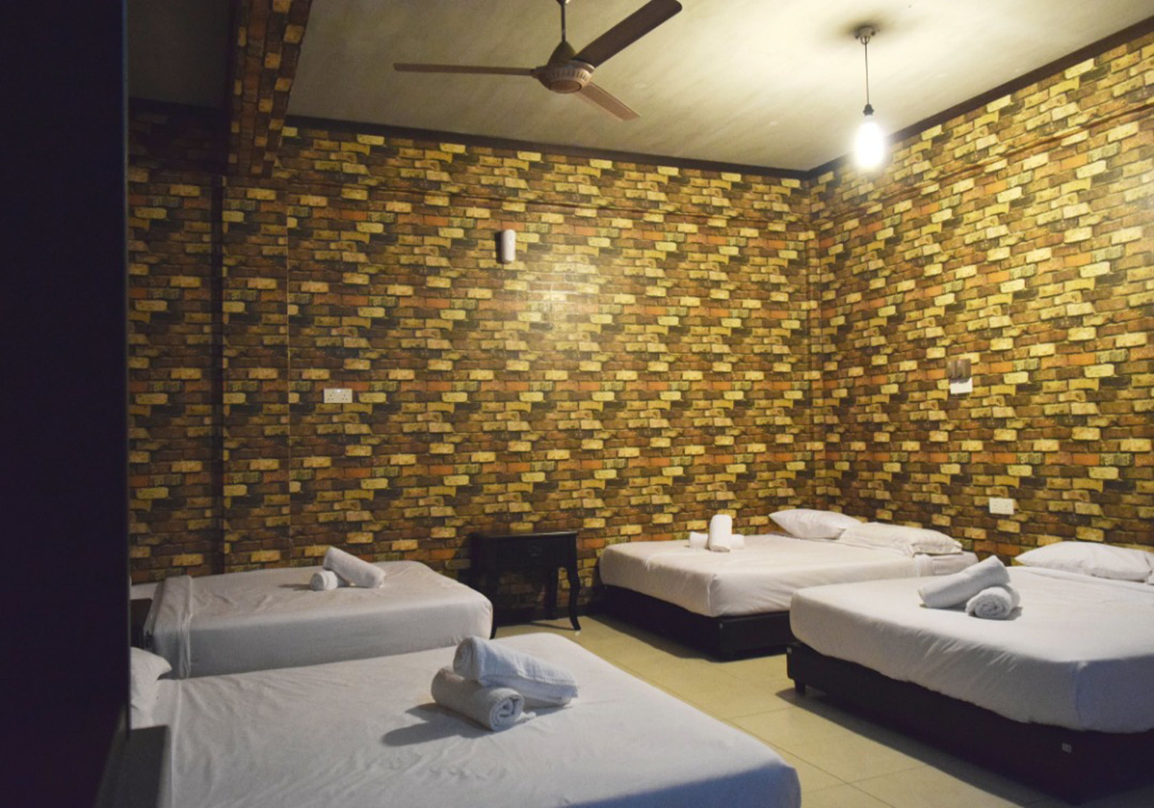Habitación de hotel con cuatro camas dispuestas en fila, un ventilador de techo y una pared con diseño de ladrillo.