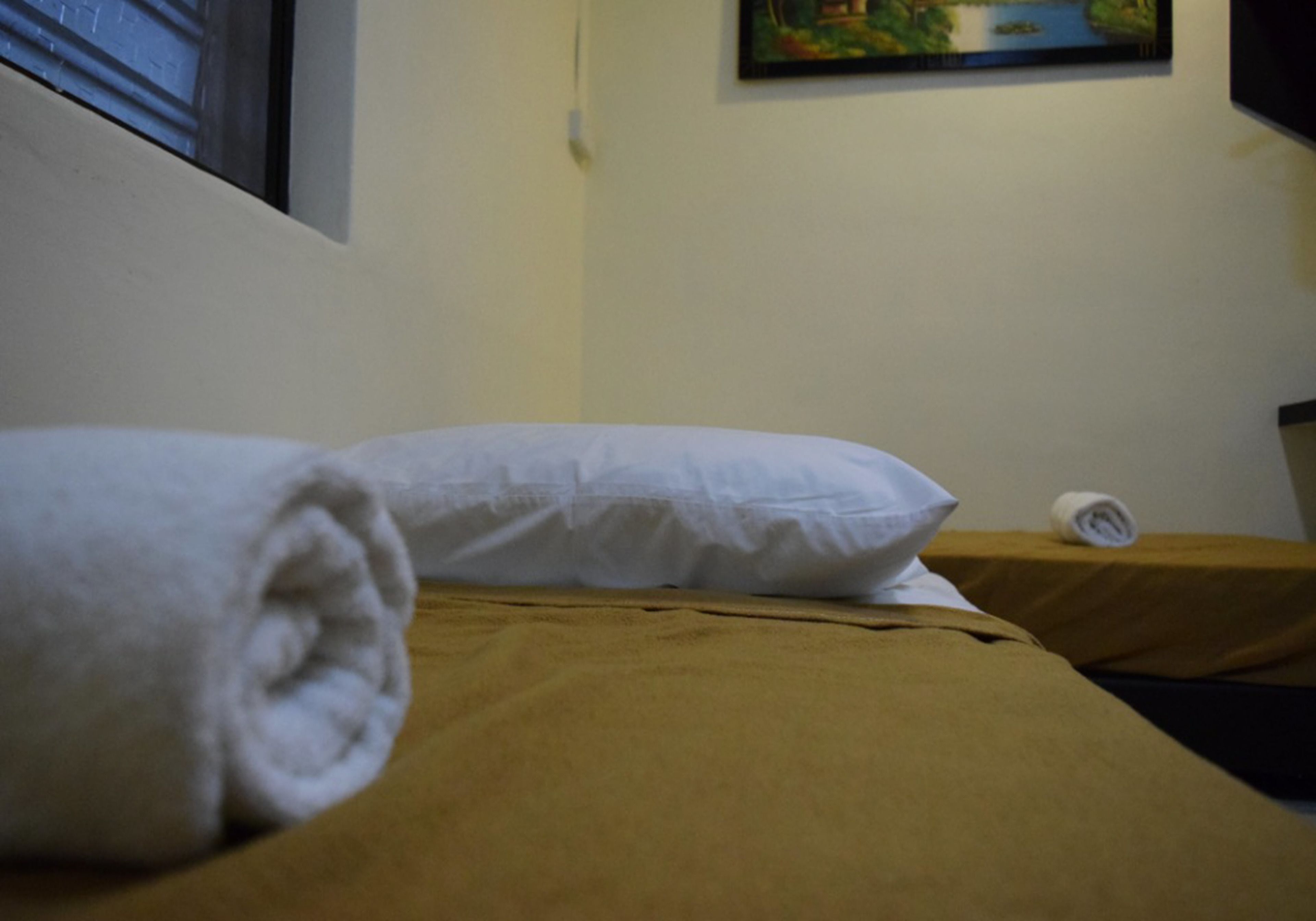 A bed with a rolled white towel and a white pillow, a brown bedspread, and a small table with a rolled white towel.