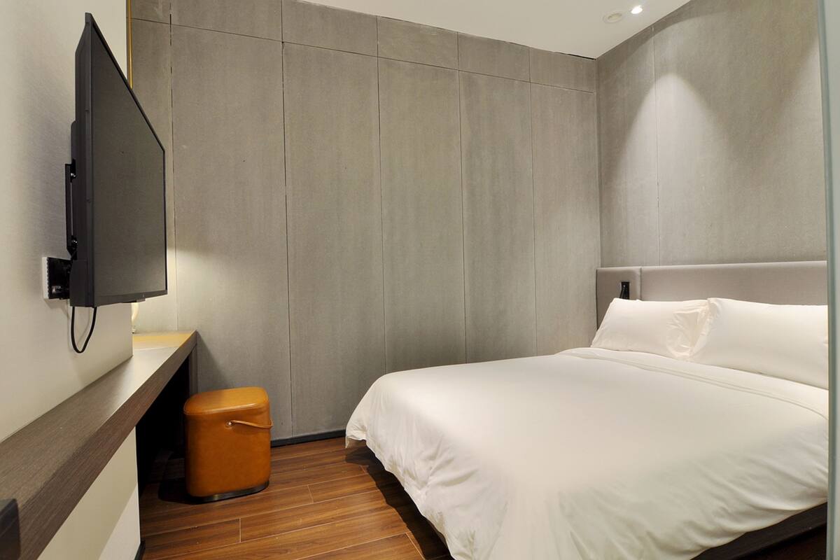 Ein modernes Hotelzimmer mit Holzboden, einem großen Bett mit weißer Bettwäsche, einem kleinen orangefarbenen Hocker, einem an der Wand befestigten Flachbildfernseher und einem gepolsterten, grauen Kopfteil.