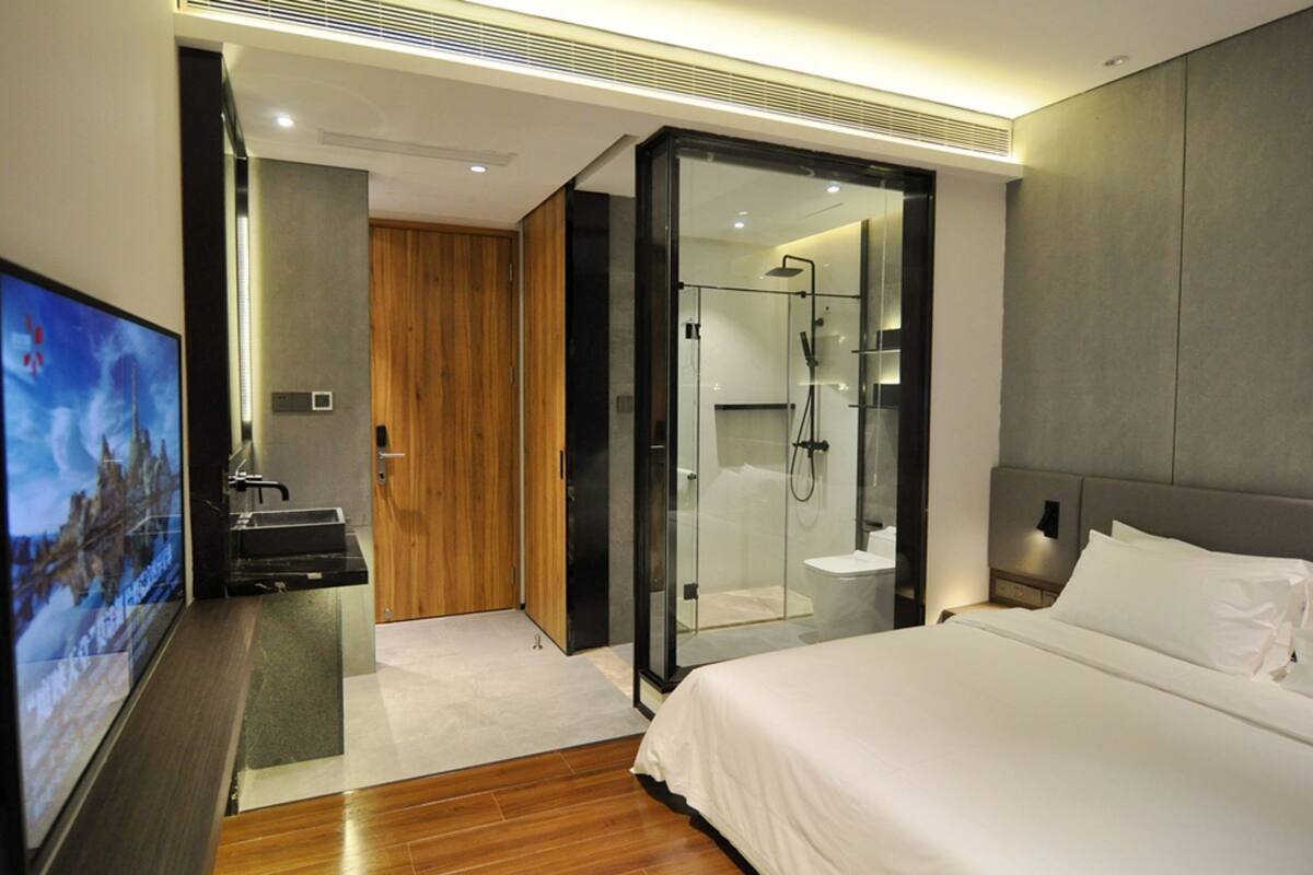 Ein modernes Hotelzimmer mit einem großen Bett, einem Fernseher, einem Holzschrank, einer Glasdusche und einem kleinen Badezimmer.