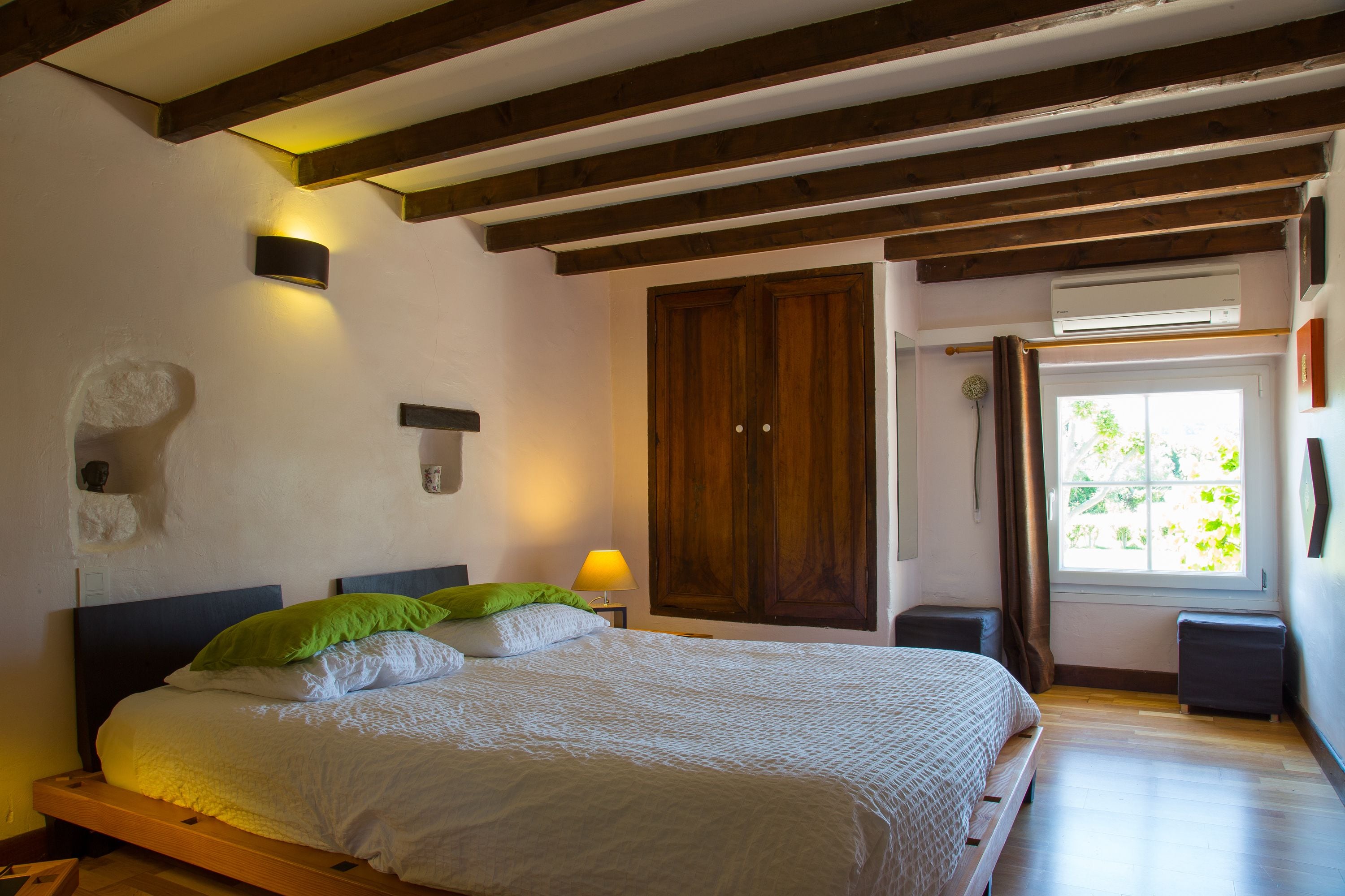 A bedroom with a wooden bed frame, a nightstand with a lamp, a wardrobe, and a window with curtains.
