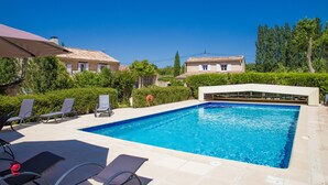 A swimming pool with lounge chairs and umbrellas, surrounded by greenery and traditional buildings. - Mas Julien (Orange)