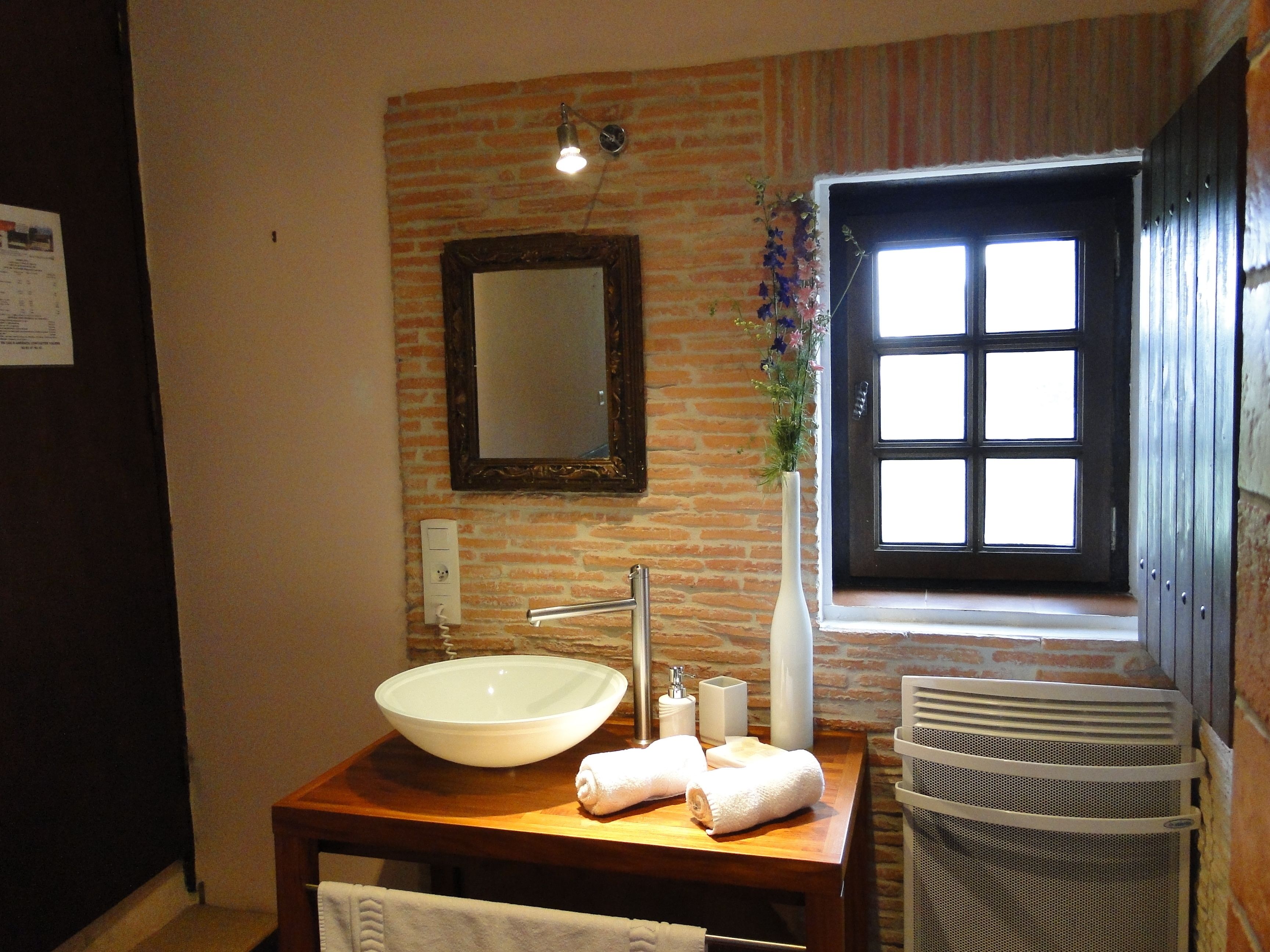 Un baño con un lavamanos grande de color blanco, un espejo encima, una ventana con persianas y un cesto de basura.