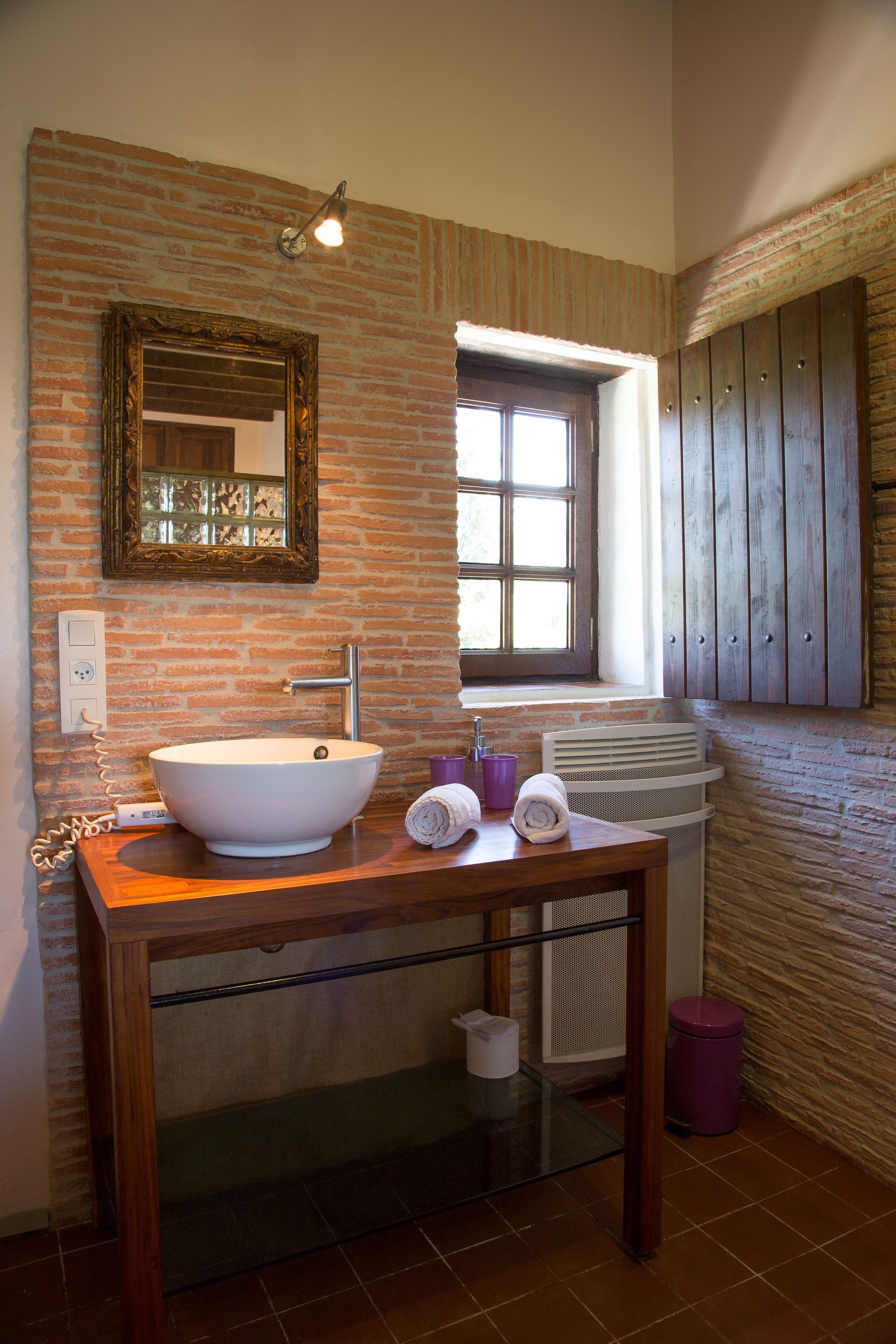 Une salle de bain avec un grand lavabo blanc, un miroir et une fenêtre. On trouve un meuble en bois au-dessus du lavabo et un porte-serviettes à côté.