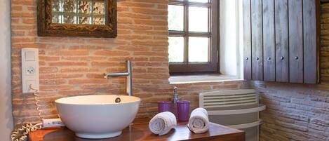 Une salle de bain avec un grand lavabo blanc, un miroir et une fenêtre. On trouve un meuble en bois au-dessus du lavabo et un porte-serviettes à côté.