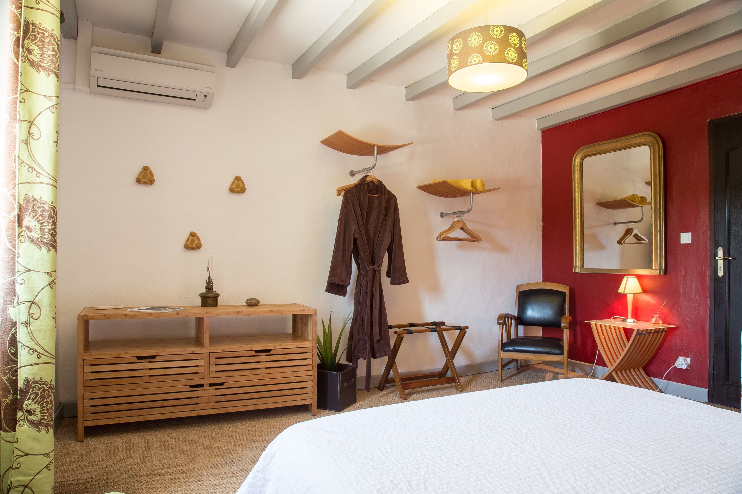 A bedroom with a bed, a wooden dresser, a chair, a robe, a mirror, and a lamp.