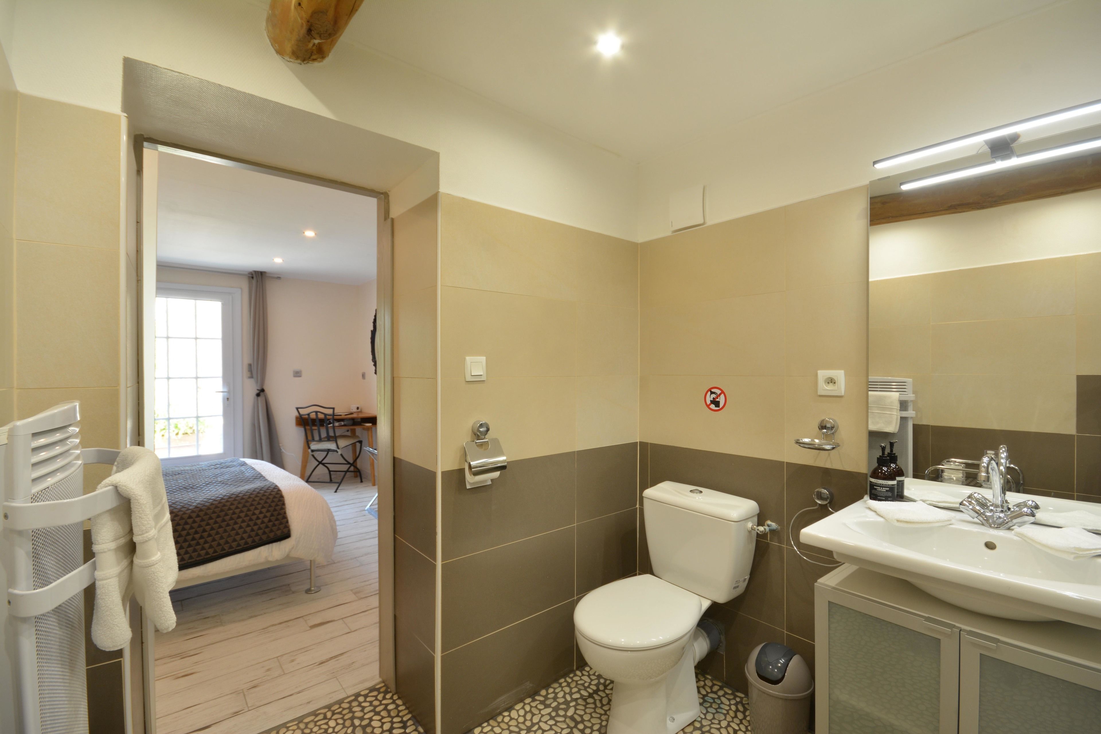 A bathroom with a toilet, sink, and shower area. There is a bed in the adjacent room.