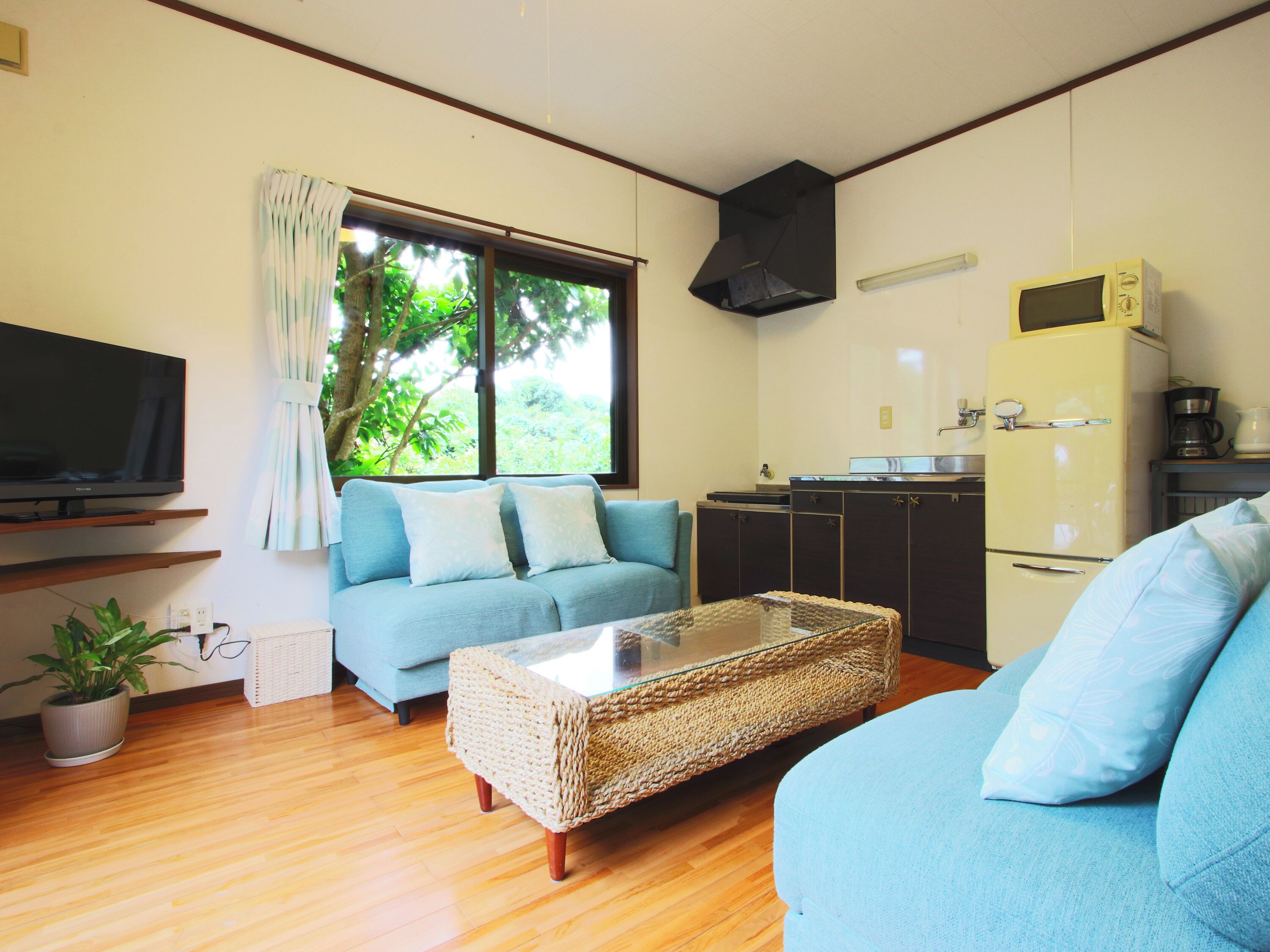 A living room with a sofa, a coffee table, a television, a kitchen area with a refrigerator, and a window with curtains.