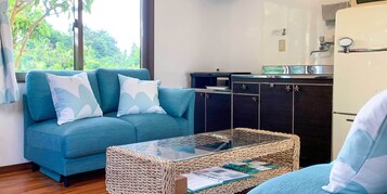 A living room with a sofa, a glass-top coffee table, and a kitchen area with cabinets and a refrigerator.