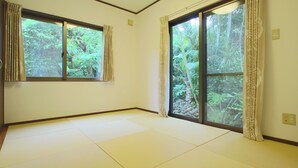 A room with tatami flooring, large windows with curtains, and a view of a forest.