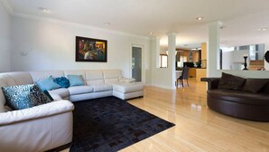 Un salon spacieux avec un canapé blanc, un tapis noir et un fauteuil en cuir marron. Un tableau est accroché au mur, et on aperçoit une cuisine au fond.