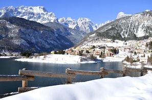Un pueblo nevado al pie de una montaña, con un lago, valla de madera y edificios con tejados rojos.