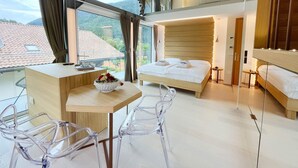 A modern hotel room with a large bed, a desk with a chair, a small table with a bowl of flowers, and a view of the mountains through large windows. - Alle Dolomiti Boutique Lake Hotel (Molveno)