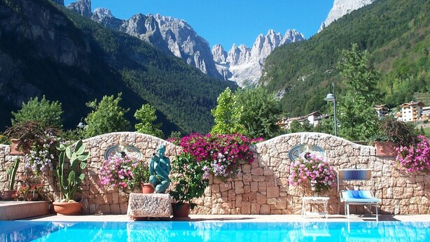 A swimming pool with a stone wall and potted plants, surrounded by mountains and houses. - Alle Dolomiti Boutique Lake Hotel (Molveno)