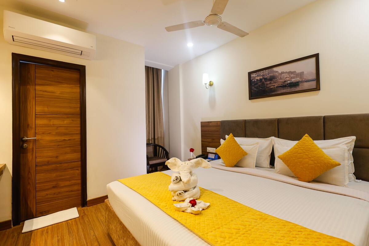 Habitación de hotel con cama, puerta de madera, aire acondicionado, ventilador de techo, un cuadro enmarcado en la pared y una pequeña mesa con una silla.