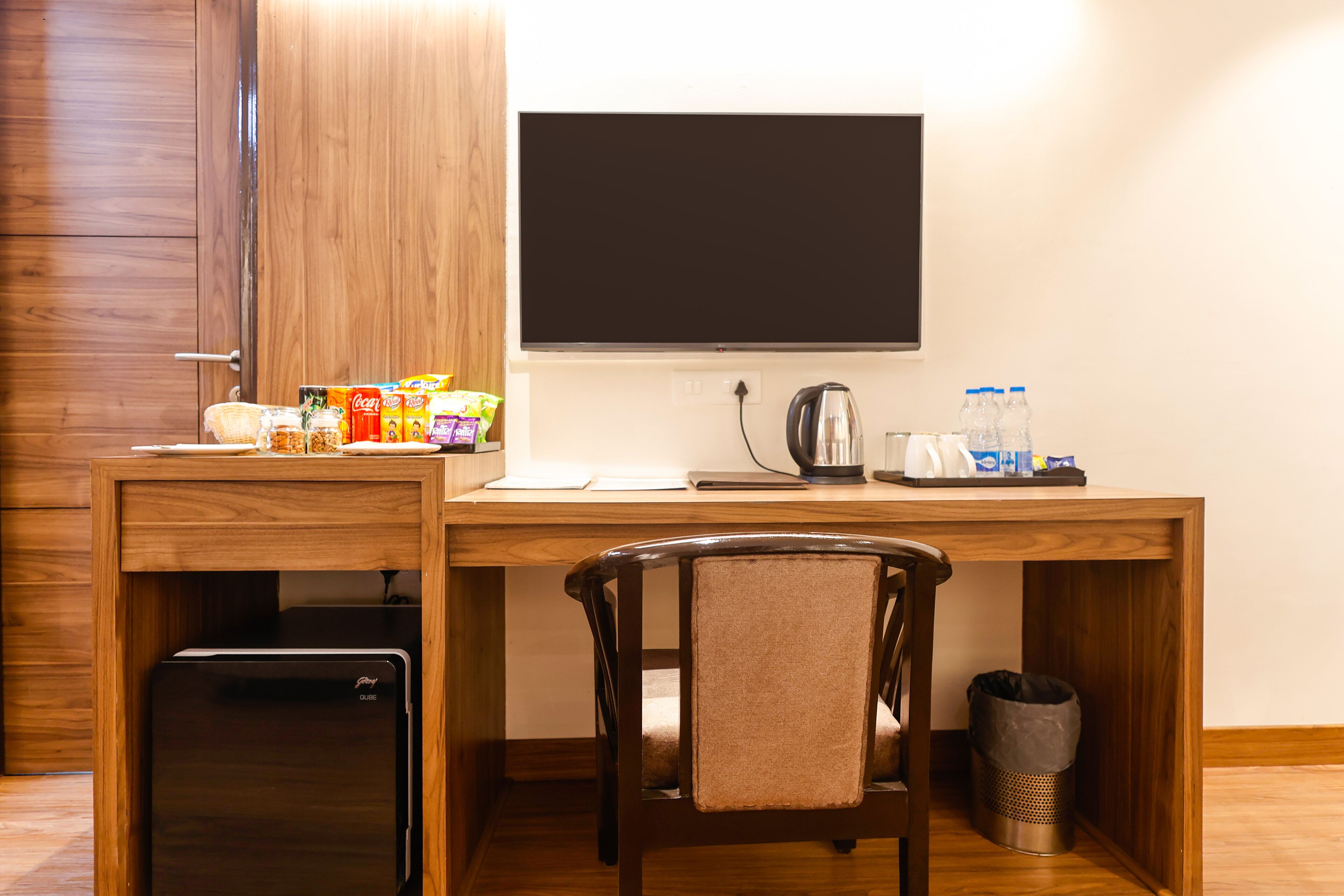 A compact hotel room with a wooden desk, a chair, a small refrigerator, a flat-screen TV, and a coffee maker.