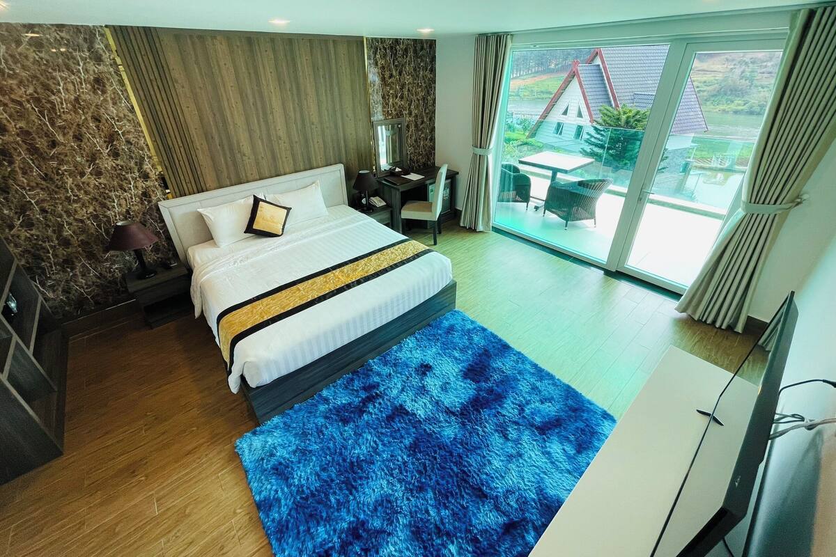 Ein modernes Hotelzimmer mit einem großen Bett, einem Schreibtisch und Blick auf ein Haus durch eine Schiebeglastür.