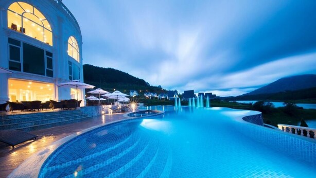 A large indoor swimming pool with a curved design, surrounded by a building with large windows and outdoor seating. - Dalat Wonder Resort (Xuan Huong - Da Lat)