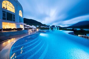 A large indoor swimming pool with a curved design, surrounded by a building with large windows and outdoor seating. - Dalat Wonder Resort (Xuan Huong - Da Lat)