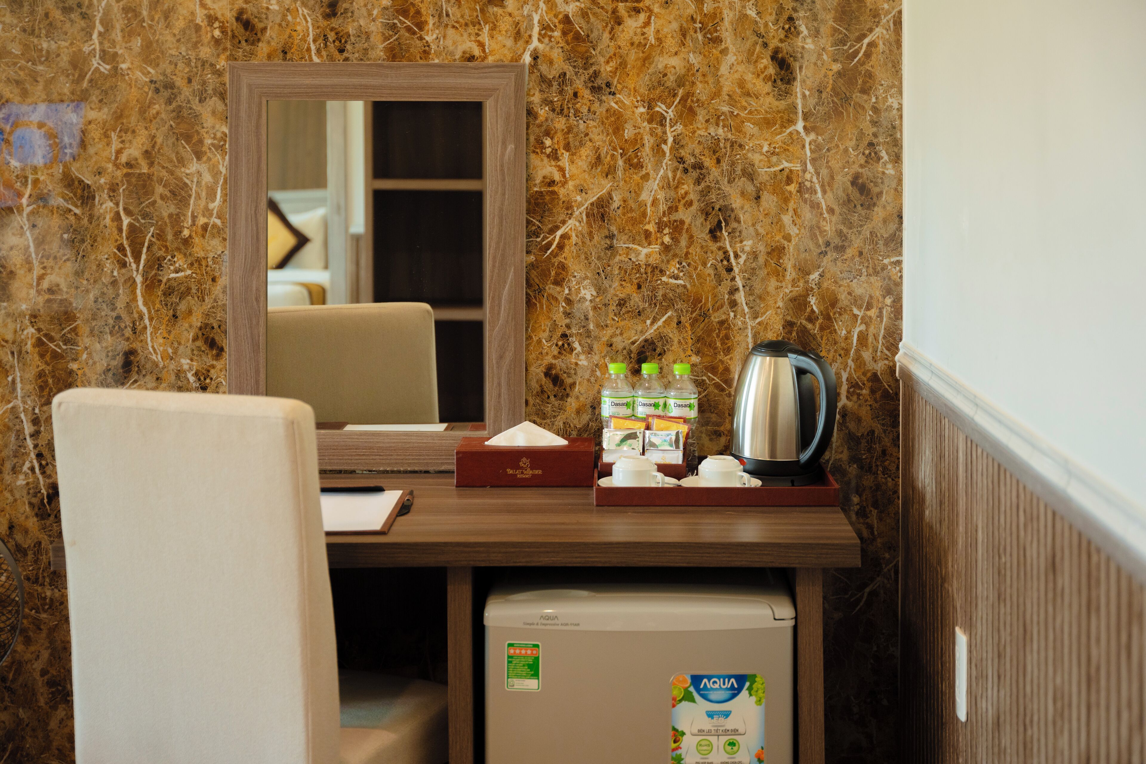 Quarto de hotel compacto com parede de mármore, pequena escrivaninha com espelho, cadeira, minibar e frigorífico.