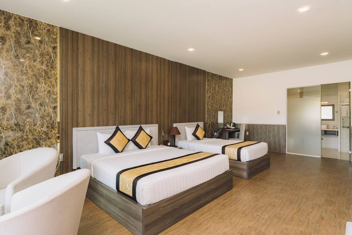 Ein Hotelzimmer mit zwei Betten, einer Sitzecke mit einem Sessel, einer Holzwand und einem durch eine Glastür einsehbaren Badezimmer.